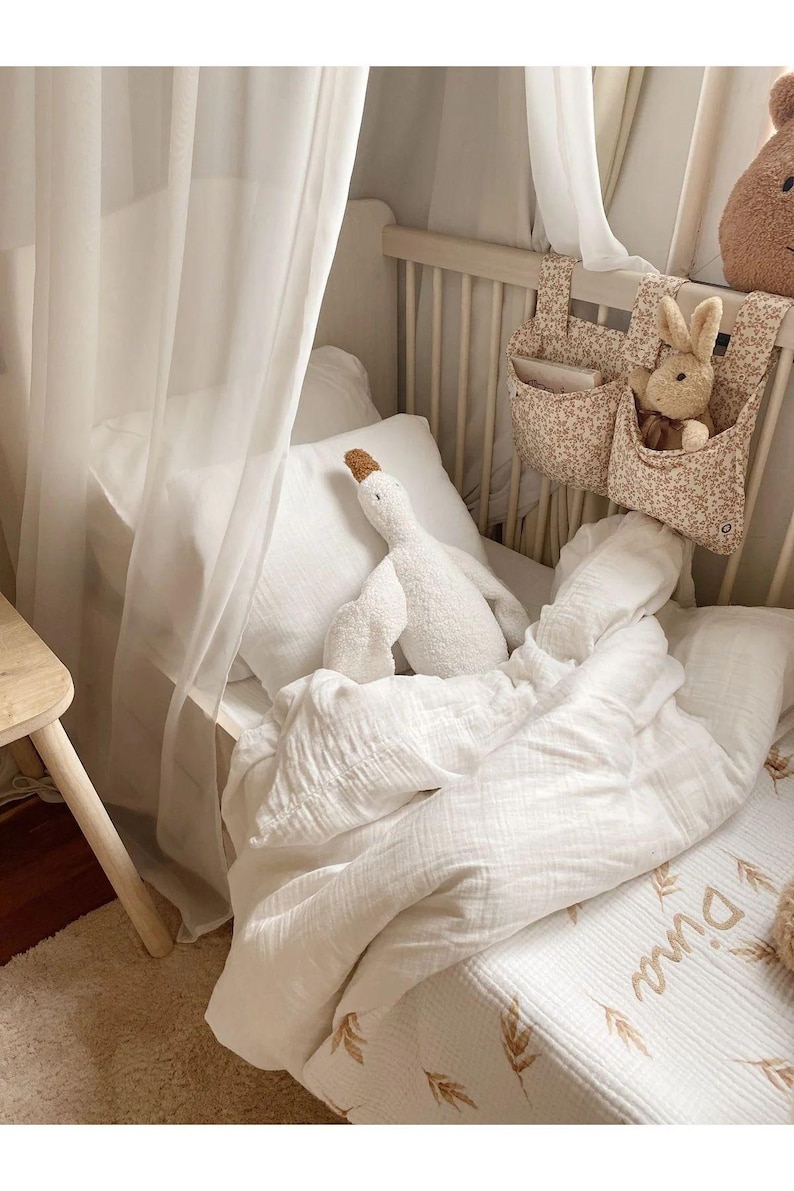 Puede incluir: Una escena de guarder&iacute;a con una cuna blanca, ropa de cama blanca y suave, y un peluche de ganso blanco. Un organizador con estampado floral contiene un conejito de peluche. La cuna est&aacute; cubierta con una cortina blanca transparente.