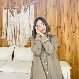 Peut inclure: Une femme porte une chemise de nuit taupe avec des bordures et des boutons blancs. La chemise de nuit a des manches longues et un col. Une tenture murale en macram&eacute; est visible en arri&egrave;re-plan.
