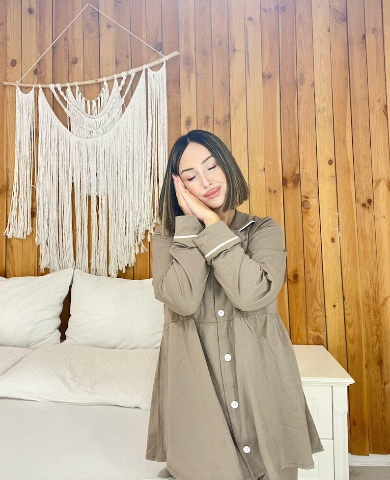 Peut inclure: Une femme porte une chemise de nuit taupe avec des bordures et des boutons blancs. La chemise de nuit a des manches longues et un col. Une tenture murale en macram&eacute; est visible en arri&egrave;re-plan.