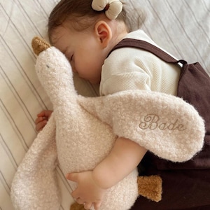 May include: A plush goose toy made of cream-coloured textured fabric with brown accents on the beak and feet. The name Bade is embroidered in cursive script on one of the wings. The toy has a soft, fuzzy appearance and a long neck design.