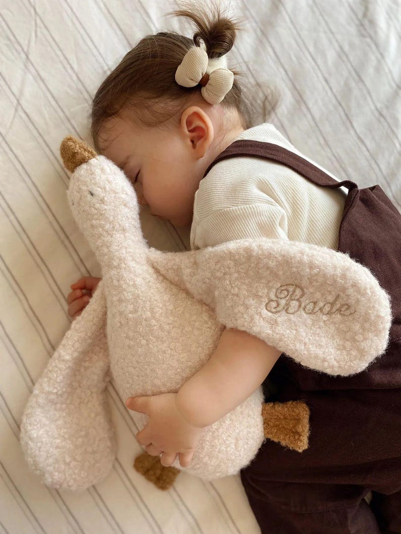 May include: A plush goose toy made of cream-coloured textured fabric with brown accents on the beak and feet. The name Bade is embroidered in cursive script on one of the wings. The toy has a soft, fuzzy appearance and a long neck design.