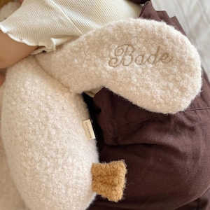 May include: A cream-coloured, textured sherpa-style plush toy shaped like a crescent moon, featuring the embroidered name "Bade" in a cursive script. A small tan fabric tag labelled "BOHEMOON" is attached to the side, and a small brown accent piece.