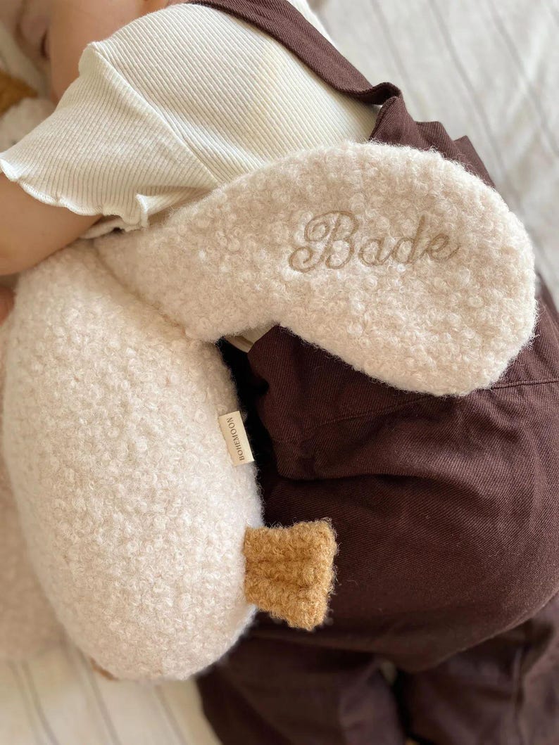 May include: A cream-coloured, textured sherpa-style plush toy shaped like a crescent moon, featuring the embroidered name "Bade" in a cursive script. A small tan fabric tag labelled "BOHEMOON" is attached to the side, and a small brown accent piece.