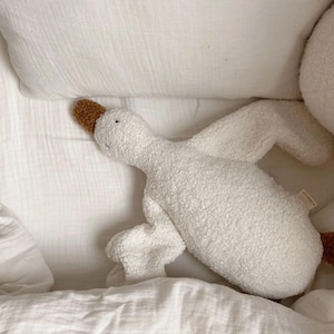 Puede incluir: Un juguete de peluche blanco con forma de ganso, con pico y patas marrones. El peluche est&aacute; tumbado en una cama blanca con una almohada y una manta blancas. El ganso tiene una textura suave y un dise&ntilde;o sencillo.