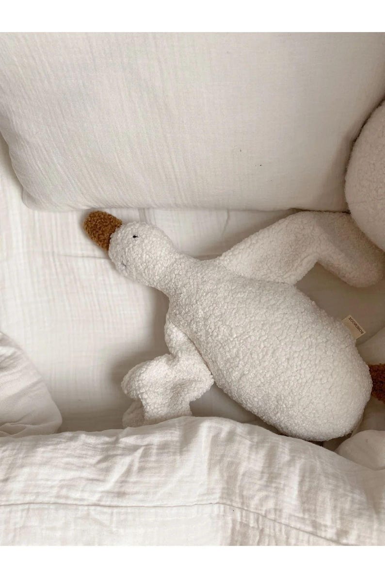 Puede incluir: Un juguete de peluche blanco con forma de ganso, con pico y patas marrones. El peluche est&aacute; tumbado en una cama blanca con una almohada y una manta blancas. El ganso tiene una textura suave y un dise&ntilde;o sencillo.