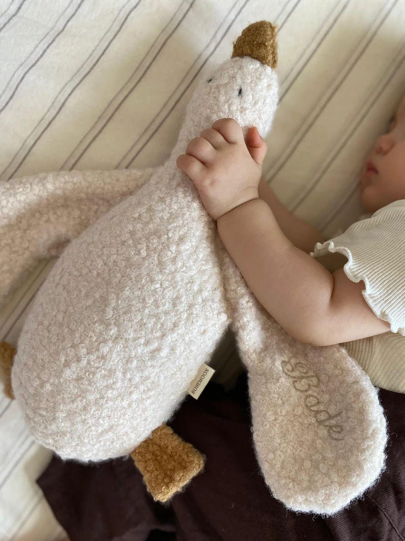 May include: A plush goose toy made of cream-coloured textured fabric with brown accents on the beak and feet. The toy features a small white tag labelled BOHEMOON and the name Bode embroidered in cursive script on one of the long, soft fabric wings.