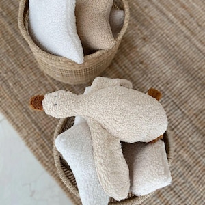 Puede incluir: Dos cestas tejidas llenas de juguetes de peluche. Una cesta contiene almohadas en forma de estrella en blanco y beige. La otra cesta contiene un juguete de ganso de peluche con pico y patas marrones, junto con almohadas en forma de estrella.