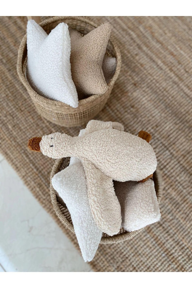 Puede incluir: Dos cestas tejidas llenas de juguetes de peluche. Una cesta contiene almohadas en forma de estrella en blanco y beige. La otra cesta contiene un juguete de ganso de peluche con pico y patas marrones, junto con almohadas en forma de estrella.