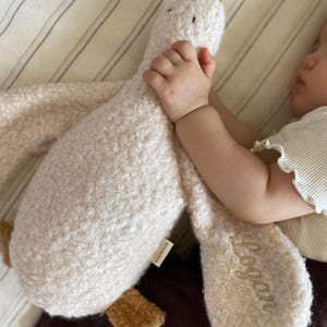 May include: A plush goose toy made of cream-coloured textured fabric with brown accents on the beak and feet. The toy features the name Logan embroidered in cursive script on one of its wings and includes a small white brand tag on the side.