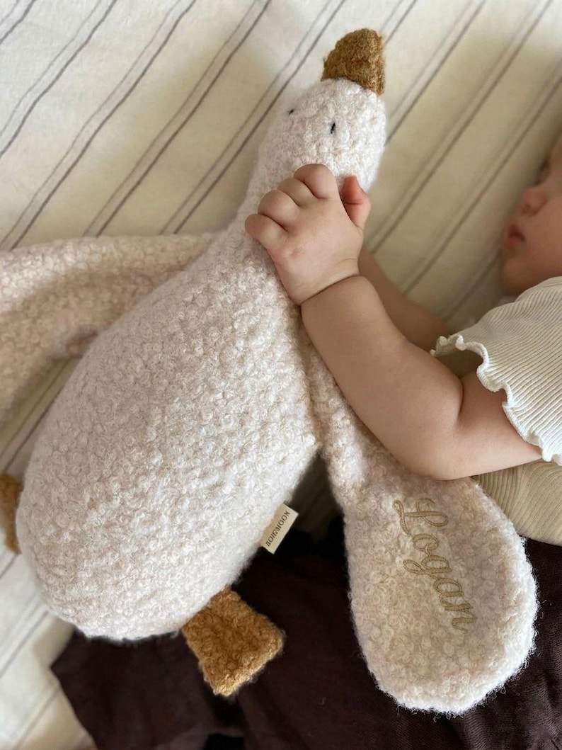 May include: A plush goose toy made of cream-coloured textured fabric with brown accents on the beak and feet. The toy features the name Logan embroidered in cursive script on one of its wings and includes a small white brand tag on the side.