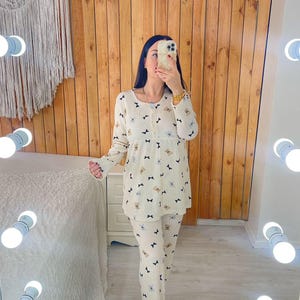 Puede incluir: Conjunto de pijama color crema con un estampado repetido de lazos negros y caras de ositos de peluche color canela. La parte superior de manga larga tiene una parte delantera abotonada y los pantalones son largos. El conjunto se muestra frente a una pared de madera y un espejo iluminado.