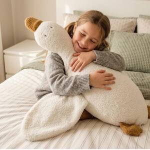 May include: A large, soft plush toy shaped like a goose, featuring a cream-colored, textured, fuzzy body with a long neck, a rounded head, and brown fabric accents on the beak and feet. The goose has simple embroidered eyes and a plush texture.