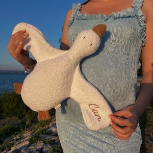 May include: A white plush toy shaped like a flying goose with a textured, fuzzy surface. The goose has brown fabric feet and a brown beak. The name "Can" is embroidered in brown cursive script on one of the wings. The toy is soft and stuffed.