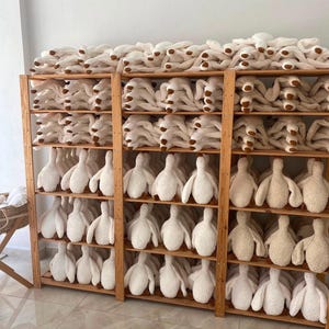 May include: A wooden shelving unit filled with rows of white plush stuffed animals shaped like geese. Each plush toy features a soft, textured fabric body with brown accents on the feet. The shelves are organised with multiple identical items.