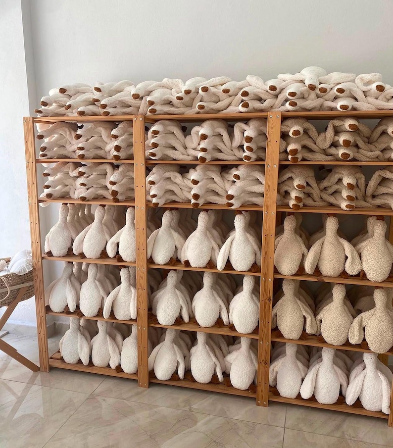May include: A wooden shelving unit filled with rows of white plush stuffed animals shaped like geese. Each plush toy features a soft, textured fabric body with brown accents on the feet. The shelves are organised with multiple identical items.