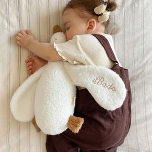 May include: A white plush goose toy with a textured, sherpa-like fabric and brown feet. The name Bade is embroidered in brown script on one of the long, floppy wings. The toy is held by a child wearing a cream-coloured shirt and brown overalls.