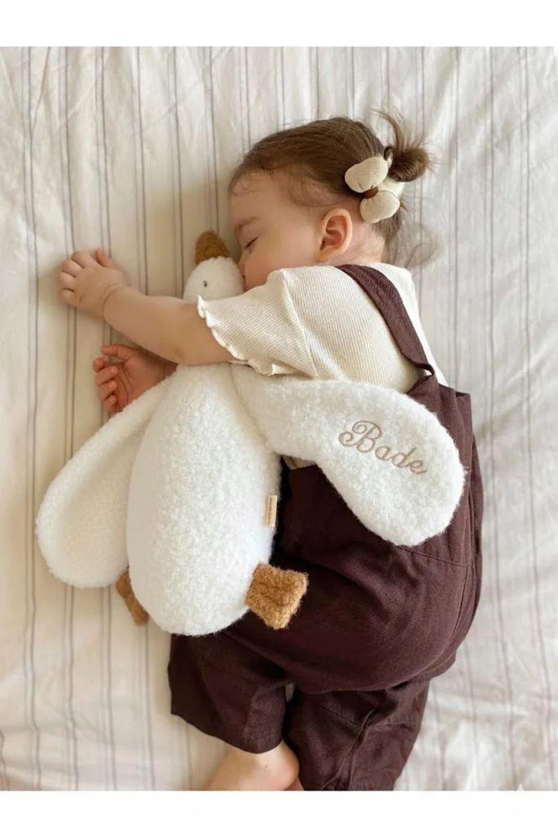May include: A white plush goose toy with a textured, sherpa-like fabric and brown feet. The name Bade is embroidered in brown script on one of the long, floppy wings. The toy is held by a child wearing a cream-coloured shirt and brown overalls.