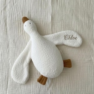 May include: A white plush goose toy with a textured, sherpa-like fabric. The goose features a brown beak and brown feet, with the name Chloe embroidered in a cursive, dark-coloured script on one of its outstretched wings.