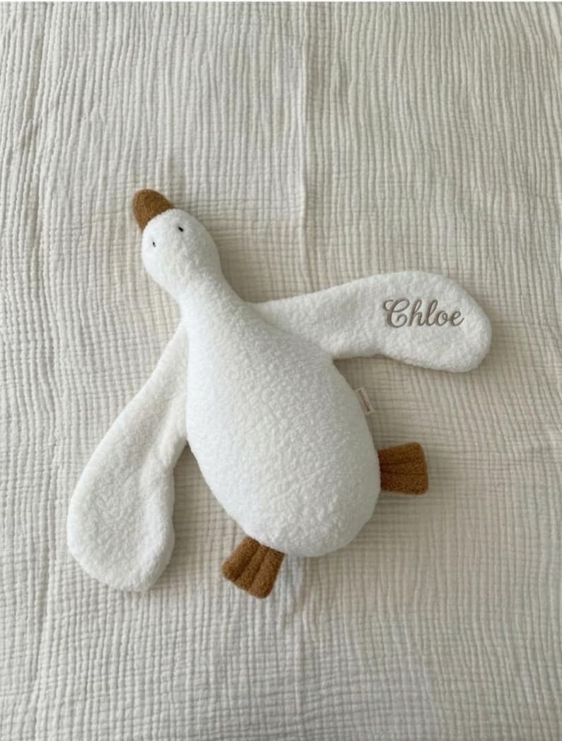 May include: A white plush goose toy with a textured, sherpa-like fabric. The goose features a brown beak and brown feet, with the name Chloe embroidered in a cursive, dark-coloured script on one of its outstretched wings.