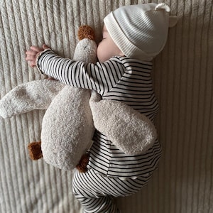Puede incluir: Un beb&eacute; dormido con gorro blanco y un body a rayas blancas y negras, abrazando un peluche blanco con detalles marrones. El beb&eacute; est&aacute; acostado sobre una superficie texturizada blanquecina.