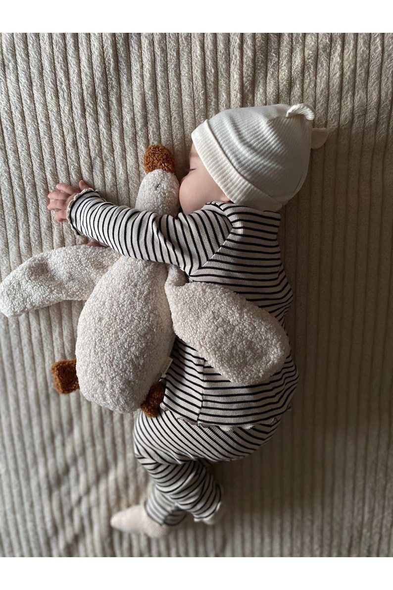 Puede incluir: Un beb&eacute; dormido con gorro blanco y un body a rayas blancas y negras, abrazando un peluche blanco con detalles marrones. El beb&eacute; est&aacute; acostado sobre una superficie texturizada blanquecina.