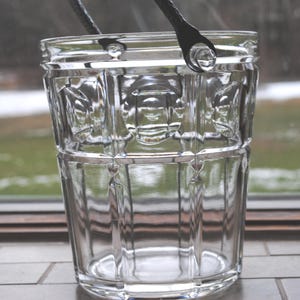 Puede incluir: Un cubo de hielo de vidrio transparente con un patrón decorativo. El cubo tiene un asa de metal con dos puntas.