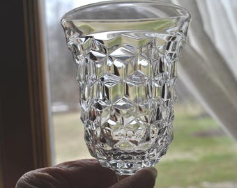 May include: A clear glass goblet featuring a prominent geometric, faceted diamond pattern across its exterior surface. The vessel has a wide, circular rim that tapers slightly toward the base, showcasing a reflective, textured design throughout.