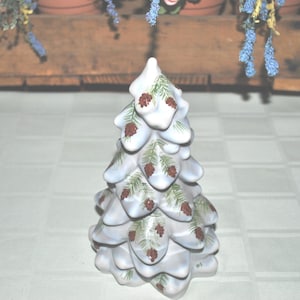Op de afbeelding: Een keramische kerstboomfiguur. De boom is wit geschilderd met blauwe tinten en heeft bruine dennenappels en groene dennennaalden. De boom is een decoratief item voor de feestdagen.