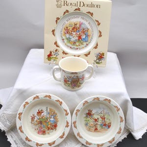 May include: A Royal Doulton Bunnykins children's dish set. The set includes a plate, two bowls, and a handled mug. Each piece features colorful illustrations of rabbits and the text "Happy Birthday From Bunnykins".