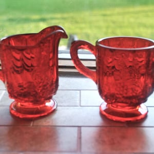 May include: A red glass creamer and sugar bowl set with a textured floral design. The creamer has a handle and a spout, while the sugar bowl has two handles.