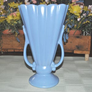 May include: A pale blue ceramic vase with a fluted rim and two curved handles. The vase has a wide, rounded base and stands approximately 25 cm tall. The vase is on a white tablecloth.