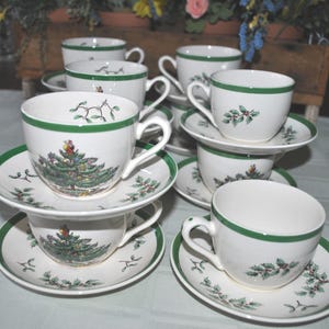 May include: A set of white teacups and saucers with green trim and festive tree and holly designs. The teacups are stacked, showcasing the full set. The saucers have a matching green border.