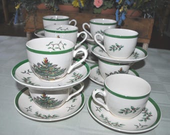 May include: A set of white teacups and saucers with green trim and festive tree and holly designs. The teacups are stacked, showcasing the full set. The saucers have a matching green border.