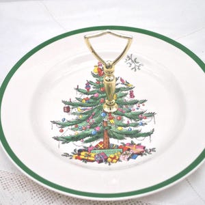 May include: A two-tiered serving plate with a Christmas tree design. The white plate has a green border and a gold handle. The tree is decorated with ornaments and presents. A festive holiday serving piece.