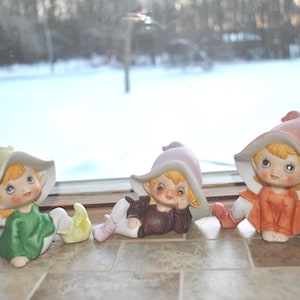 May include: Three ceramic elf figurines with large hats and sweet faces. One elf wears a green outfit and a yellow hat, another wears a purple hat and brown outfit, and the third wears an orange outfit and a pink hat. The figurines are on a tiled surface.