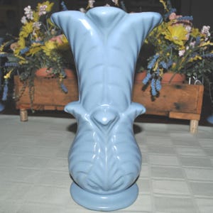 May include: A pale blue ceramic vase with a flared top and a rounded base. The vase features a detailed, leaf-like design and stands approximately 25 cm tall. The vase is on a white tablecloth.