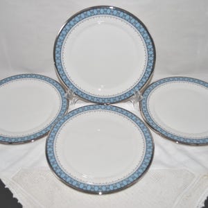 May include: A set of four round white ceramic plates featuring a decorative border pattern in light blue and dark blue with a silver metallic rim. The plates have a smooth, glossy finish and a consistent circular design across all four pieces.
