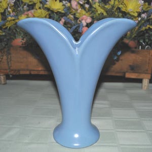 May include: A light blue ceramic vase with a distinctive, flared design. The vase has a rounded base and two upward-curving, petal-like openings. The vase is approximately 25 cm tall and is set against a background of flowers.
