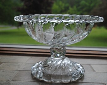 May include: A clear glass pedestal bowl with a swirl design. The bowl has a wide, shallow base and a tall, slender stem.