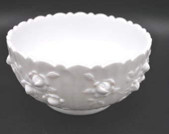 May include: A white ceramic bowl with a scalloped rim and embossed rose detailing. The bowl has a round base and a smooth interior. The exterior features several raised rose designs. The bowl is set against a dark background.