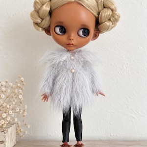 Könnte beinhalten: Eine Puppe mit gebräunter Haut und großen dunklen Augen, deren blondes Haar zu zwei geflochtenen Dutts frisiert ist. Die Puppe trägt einen langärmeligen, hellgrauen Kunstpelzmantel mit zwei kleinen perlmuttartigen Knöpfen, dazu eine schwarze Leggings und braune Schnürschuhe.