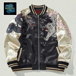 Pode incluir: Casaco bomber de cetim preto com mangas creme. Apresenta bordados de dragão e tigre, com detalhes de flores de cerejeira. O casaco tem fecho de correr, punhos listrados e o texto "DAGODDCHANCE BY I.C." num logotipo floral azul.