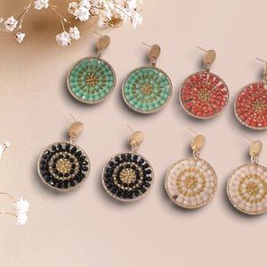 Puede incluir: Una colección de pendientes colgantes redondos en varios colores, incluyendo turquesa, rojo, negro y blanco. Cada pendiente presenta un marco y un poste dorados, con un diseño de cuentas en el centro. Los pendientes se muestran sobre un fondo rosa claro.