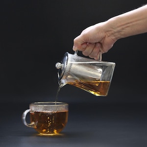 Puede incluir: Una tetera de vidrio transparente con un infusor de acero inoxidable vierte té de color ámbar en una taza de té de vidrio transparente. La tetera tiene un asa y una tapa. La taza tiene un asa.