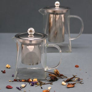 Pode incluir: Duas chaleiras de vidro transparente com tampas e infusores de aço inoxidável. As chaleiras têm bases quadradas e alças curvas. Folhas de chá e pétalas de flores secas estão espalhadas.