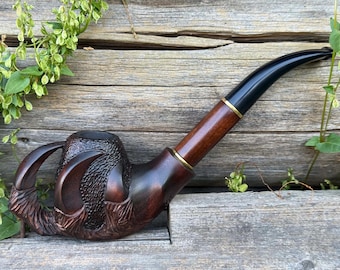 May include: A dark brown wooden tobacco pipe featuring a carved claw design on the bowl. The pipe has a smooth, polished finish, a long curved black stem, and a thin gold-colored metal band connecting the wooden bowl to the black mouthpiece.