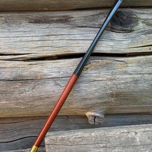 Puede incluir: Una boquilla para cigarrillos larga y delgada que presenta un cuerpo de madera de color marrón rojizo liso, una boquilla negra brillante y una pequeña banda de metal dorado pulido que conecta las dos secciones. El objeto es recto y se estrecha.