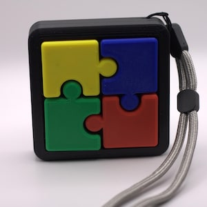May include: A black square puzzle with four colorful pieces: yellow, blue, green, and red. The puzzle has a wrist strap attached. The pieces are in a jigsaw puzzle design.