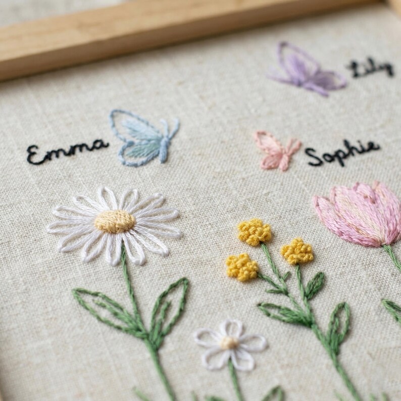 Puede incluir: Obra de arte bordada con una variedad de flores y mariposas coloridas. Los nombres "Emma", "Sophie" y "Lily" est&aacute;n bordados encima de las flores y mariposas. La obra est&aacute; enmarcada en un marco de madera clara.