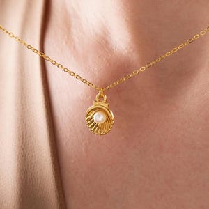 Könnte beinhalten: Eine goldfarbene Halskette mit einem kleinen Muschel-Anhänger. Der Anhänger hat eine Perle in der Mitte und hängt an einer zarten Kette. Die Halskette wird vor einem neutralen Hintergrund präsentiert.